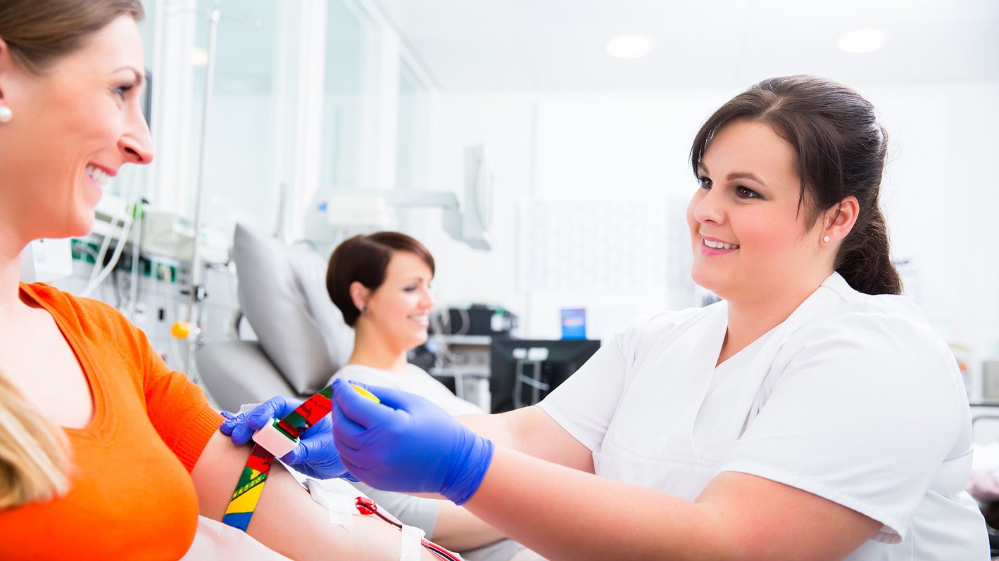 Image of a phlebotomy technician smiling while a patient sits in the background.
